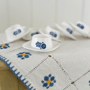 Könnte beinhalten: Eine Sammlung weißer Keramik-Eierbecher mit blauen Blumenmustern, jeweils auf einer passenden Untertasse. Die Tassen und Untertassen sind auf einer weißen, bestickten Tischdecke mit blauen und gelben Blumenmustern angeordnet.