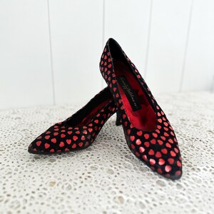 Könnte beinhalten: Ein Paar schwarze High Heels mit einem Muster aus roten Herzen. Die Schuhe haben spitze Zehen und einen kleinen Absatz. Die Innenseite des Schuhs ist rot. Die Schuhe stehen auf einer weißen Spitzentischdecke.