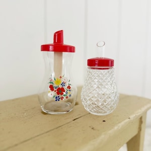 Könnte beinhalten: Zwei Zuckerspender aus Glas mit roten Deckeln. Einer hat ein Blumendesign mit roten, gelben und blauen Blumen. Der andere hat ein Rautenmuster. Beide stehen auf einer hellen Holzoberfläche.