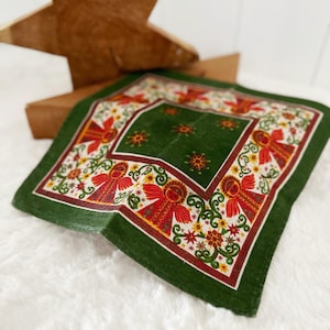 May include: A green and white Christmas-themed handkerchief with a decorative border featuring red angels, yellow flowers, and green vines. The center of the handkerchief is a solid green color with yellow starburst designs. The handkerchief is square-shaped.