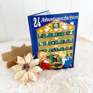 Könnte beinhalten: Ein blaues Hardcover-Buch mit dem Titel 24 Adventsgeschichten, das ein goldenes Haus mit vierundzwanzig nummerierten Fenstern, zwei Engeln und zwei Kindern zeigt. Eine cremefarbene Papierblume mit kleinen Zapfen und roten Beeren liegt daneben.