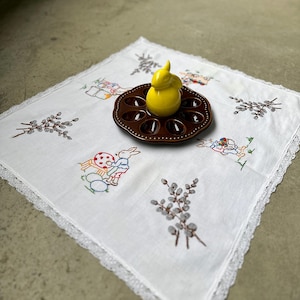 Könnte beinhalten: Ein weißes Ostertischtuch mit gestickten Hasen, Eiern und Weidenkätzchenzweigen. Ein brauner Keramik-Eierhalter mit einer gelben Hasenfigur liegt auf dem Tuch. Das Tischtuch hat eine Spitzenborte.