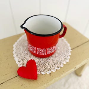 Könnte beinhalten: Eine rote Emaille-Kanne mit schwarzem Rand und Henkel, verziert mit einem weißen Zierband. Ein kleines rotes Herz liegt neben der Kanne auf einer weißen Spitzendecke. Die Kanne steht auf einer hellen Holzoberfläche.