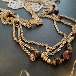 May include: Gold-toned necklace with multiple strands and a clasp. The necklace features dark red and gold beads, along with smaller silver-toned beads. The necklace is displayed on a dark surface with a white lace doily in the background. Jewelry.