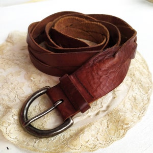 May include: A brown leather belt with a silver-colored buckle. The belt is made of braided leather and has a textured appearance. The buckle is a simple, half-moon design. The belt is shown coiled on a cream-colored lace doily.