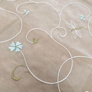May include: A sheer beige fabric featuring intricate white embroidered swirling vines, light blue flowers, and gold-toned dragonflies. The delicate embroidery creates a repeating pattern across the surface, highlighting the contrast between the light base and the detailed thread work.