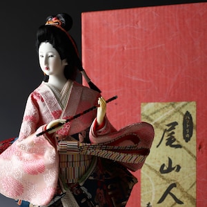 Op de afbeelding: Japanse pop in traditionele kimono, met een fluit. De pop heeft een lichte teint, zwart haar en is versierd met een rood-roze kimono met patroon. De achtergrond heeft een rode doos met Japanse tekst.