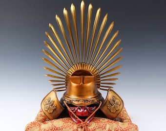 May include: A gold samurai helmet with a red face mask and a gold sunburst design on top. The helmet is sitting on a red and gold patterned cushion.