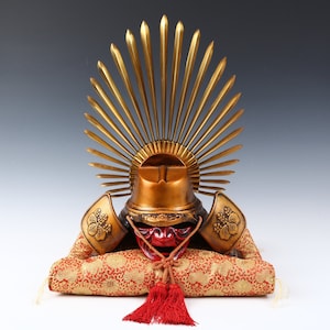 Puede incluir: Un casco de samurái dorado con una máscara facial roja y un diseño de sol dorado en la parte superior. El casco está sentado sobre un cojín con estampado rojo y dorado.