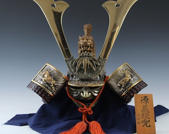 May include: An ornate Japanese samurai helmet with gold-colored horns and intricate metalwork. The helmet features a detailed face guard, decorative side panels with tiger motifs, and a wooden plaque with Japanese characters. The helmet rests on a blue cloth with orange tassels.