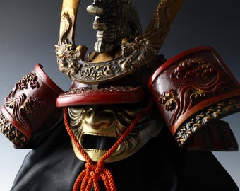 May include: Close-up of a Japanese samurai helmet. The helmet features a gold-colored face mask with an open mouth, red accents, and intricate gold and bronze detailing. A red cord hangs from the mask. The helmet is set against a dark background.