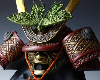 May include: Close-up of a detailed Japanese samurai helmet. The helmet features a gold-colored face mask, red and gold accents, and a green butterfly-shaped crest. The design includes intricate patterns and a black base.