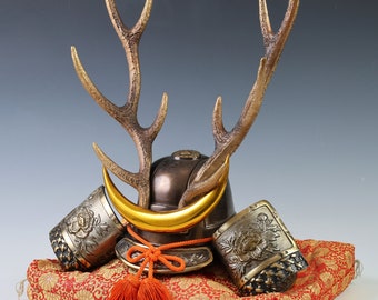May include: A miniature samurai helmet with a gold crescent moon and antlers, resting on a red and gold patterned cushion.