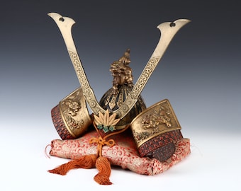 May include: A traditional Japanese samurai helmet, or kabuto, with gold and black accents, resting on a red and gold patterned cushion. The helmet features intricate details and a pair of decorative horns.