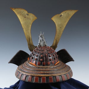 Puede incluir: Un casco de samur&aacute;i japon&eacute;s antiguo, con un dise&ntilde;o detallado. El casco tiene una combinaci&oacute;n de colores rojo oscuro y negro con acentos dorados en forma de alas. El casco est&aacute; adornado con un elemento decorativo plateado en la parte superior y detalles de cord&oacute;n naranja.