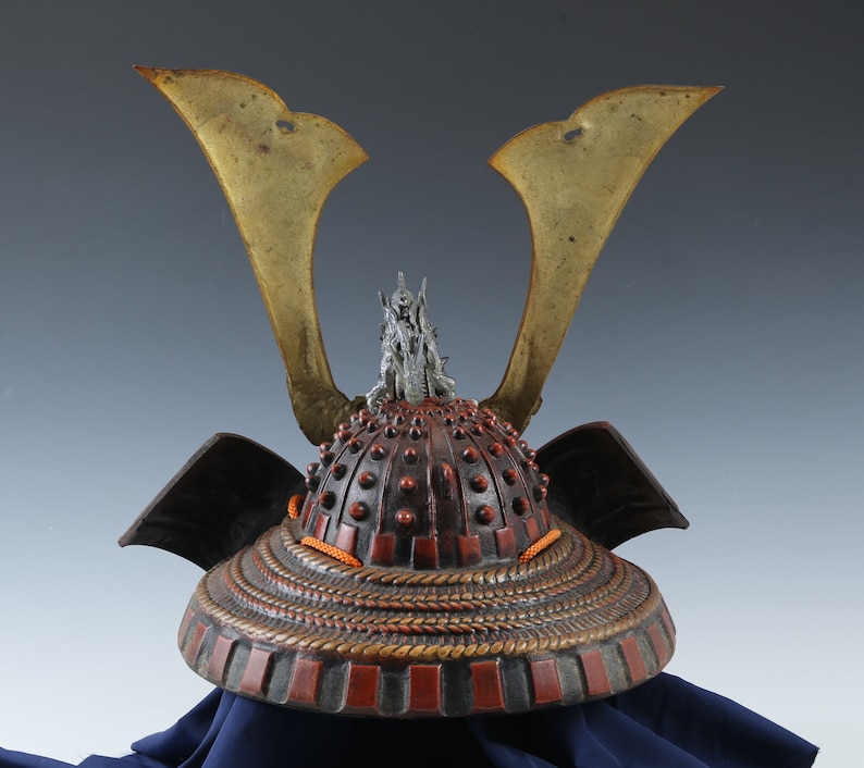 Puede incluir: Un casco de samur&aacute;i japon&eacute;s antiguo, con un dise&ntilde;o detallado. El casco tiene una combinaci&oacute;n de colores rojo oscuro y negro con acentos dorados en forma de alas. El casco est&aacute; adornado con un elemento decorativo plateado en la parte superior y detalles de cord&oacute;n naranja.