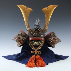 Puede incluir: Un antiguo casco de samur&aacute;i japon&eacute;s con cuernos dorados y detalles intrincados de dragones. El casco presenta una protecci&oacute;n facial y protectores de hombros rojos y bronce, con borlas naranjas colgando debajo. El casco descansa sobre una tela azul.