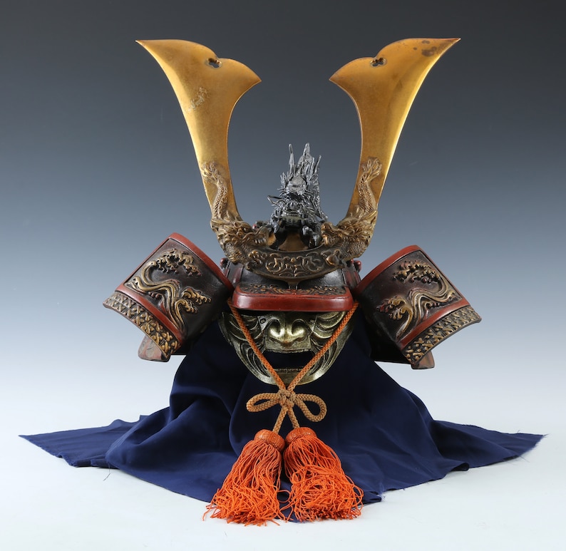 Puede incluir: Un antiguo casco de samur&aacute;i japon&eacute;s con cuernos dorados y detalles intrincados de dragones. El casco presenta una protecci&oacute;n facial y protectores de hombros rojos y bronce, con borlas naranjas colgando debajo. El casco descansa sobre una tela azul.