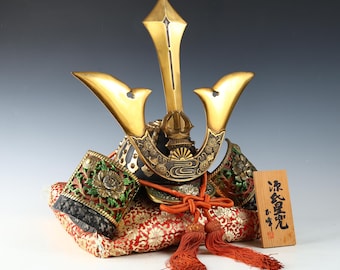 May include: A decorative Japanese samurai helmet with gold accents and intricate detailing. The helmet rests on a red patterned cushion and is accompanied by a wooden plaque with Japanese characters. The helmet features a central crest and side ornaments.
