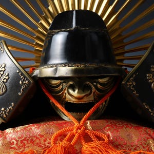 Puede incluir: Primer plano de un casco de samurái japonés. El casco negro tiene detalles dorados, incluyendo una máscara facial y elementos decorativos. El fondo presenta un diseño de rayos de sol dorados y una tela estampada en rojo y dorado.