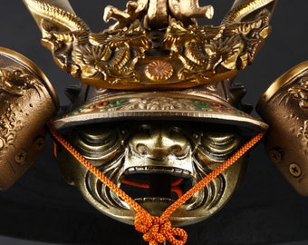 May include: Close-up of a detailed golden samurai helmet. The helmet features intricate dragon and floral designs, with a fierce face mask and orange cord. The helmet is a decorative item, likely for display or cosplay.
