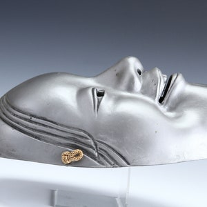 Könnte beinhalten: Eine silberne Theatermaske mit geschlossenem Auge und offenem Mund, die auf einem durchsichtigen Ständer ruht. Die Maske hat eine goldene Schnur, die um die Seite gebunden ist.