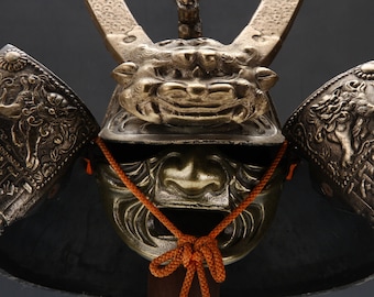 May include: A traditional Japanese samurai helmet, or kabuto, with intricate gold and silver detailing. The helmet features a fierce lion face and is adorned with a red cord.