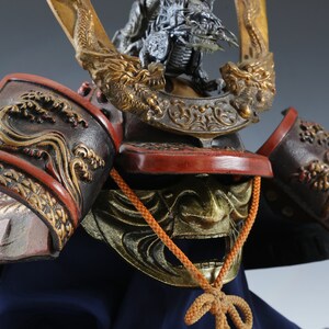 Puede incluir: Primer plano de un casco de samur&aacute;i japon&eacute;s. El casco presenta intrincados detalles dorados y rojos, incluyendo motivos de dragones y olas. Se ve una m&aacute;scara dorada y un protector de cuello, sobre una base de tela azul.