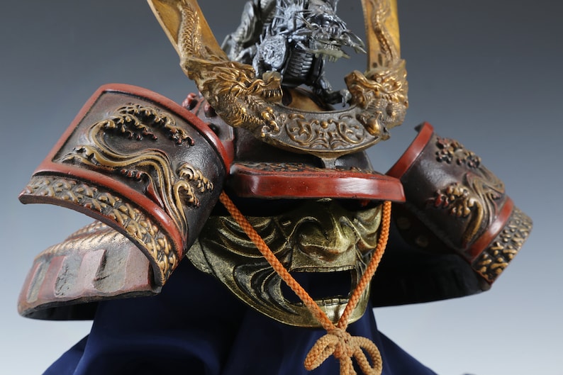 Puede incluir: Primer plano de un casco de samur&aacute;i japon&eacute;s. El casco presenta intrincados detalles dorados y rojos, incluyendo motivos de dragones y olas. Se ve una m&aacute;scara dorada y un protector de cuello, sobre una base de tela azul.