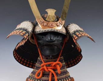 May include: A detailed view of a Japanese samurai helmet, featuring a black face mask with a mustache, gold accents, and intricate metalwork. The helmet has a neck guard and is secured with an orange cord. The overall design is ornate and traditional.