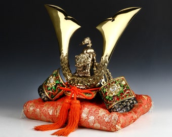 May include: A gold samurai helmet with a lion figurine on top, resting on a red and gold cushion. The helmet has intricate details and is decorated with green and red accents.