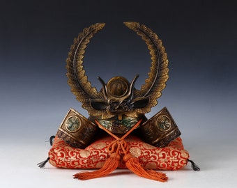 May include: A miniature samurai helmet with a gold and black finish, adorned with a gold leaf wreath. The helmet is displayed on a red velvet cushion with gold tassels.