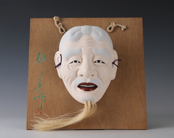 Könnte beinhalten: Eine weiße japanische Noh-Maske mit einem langen weißen Bart, bemalt mit schwarzen Augen und einem roten Mund. Die Maske ist auf einer Holztafel montiert, an der Ohren ist eine violette Schnur befestigt.