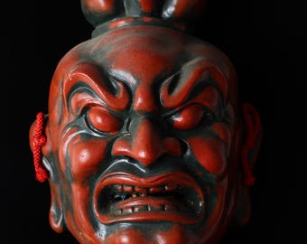 May include: A red and black Japanese Noh mask with an angry expression. The mask features a prominent brow, furrowed lines, and a snarling mouth. Red cords are attached on each side. The mask is set against a black background.