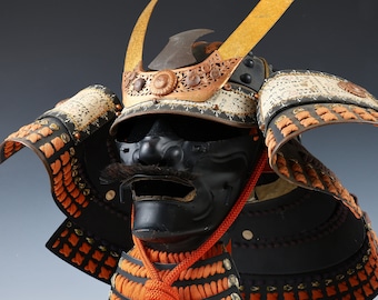 Puede incluir: Un casco de samurái negro con una máscara facial dorada y naranja y un casco dorado y blanco con un cuerno dorado. El casco está decorado con intrincados detalles y es un hermoso ejemplo de la artesanía japonesa.