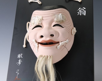 Könnte beinhalten: Eine traditionelle japanische Noh-Maske mit weißem Bart und einem lächelnden Ausdruck. Die Maske ist auf einer schwarzen Holztafel montiert, auf der die japanischen Schriftzeichen "翁" und "翁" in Weiß geschrieben sind.