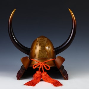 Könnte beinhalten: Ein traditioneller japanischer Samurai-Helm mit zwei großen Hörnern, einer braunen und goldenen Oberfläche und roten Quasten.
