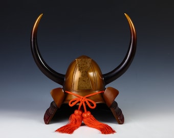 Puede incluir: Un casco de samurái japonés tradicional con dos cuernos grandes, un acabado marrón y dorado y borlas rojas.