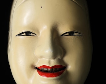 Könnte beinhalten: Eine traditionelle japanische Noh-Maske mit einem ruhigen Ausdruck. Die Maske hat einen hellen Teint, schwarz bemalte Augen und einen roten, lächelnden Mund. Das Haar ist mit schwarzen Akzenten versehen. Der Hintergrund ist tiefschwarz.