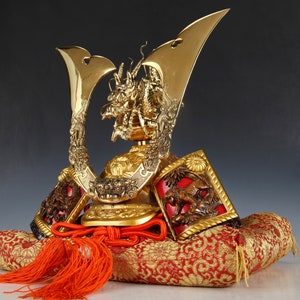 May include: A golden Japanese samurai helmet, featuring intricate dragon and floral designs. The helmet rests on a red and gold patterned cushion with an orange tassel. The helmet's details are finely crafted.