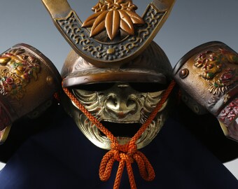 May include: A traditional Japanese samurai helmet with gold and red accents. The helmet is decorated with intricate carvings and features a fierce-looking mask.