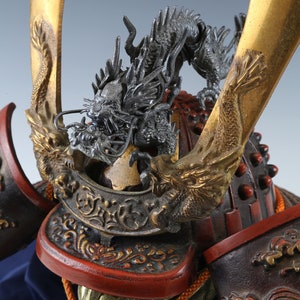Puede incluir: Primer plano de un casco de samur&aacute;i japon&eacute;s. El casco presenta un detallado escudo de drag&oacute;n plateado sobre una superficie lacada en rojo y negro. Se ven detalles dorados y elementos decorativos.