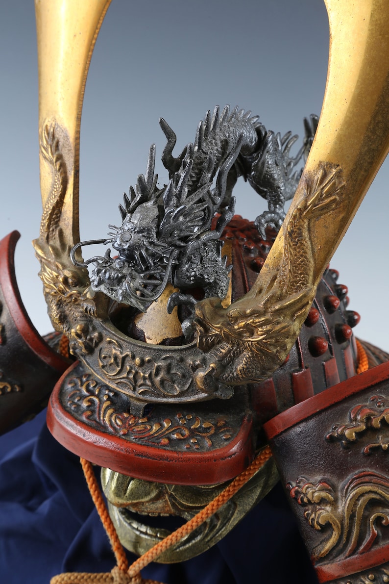 Puede incluir: Primer plano de un casco de samur&aacute;i japon&eacute;s. El casco presenta un detallado escudo de drag&oacute;n plateado sobre una superficie lacada en rojo y negro. Se ven detalles dorados y elementos decorativos.