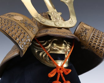 May include: A traditional Japanese samurai helmet, or kabuto, with a gold-colored face mask and intricate details. The helmet is decorated with floral patterns and has a distinctive crest.