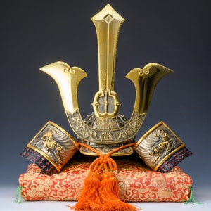 Pode incluir: Um capacete de samurai dourado com um design de tigre nos painéis laterais, descansando sobre um almofada com padrão vermelho e dourado. O capacete tem uma crista alta e pontiaguda e é decorado com detalhes intrincados.