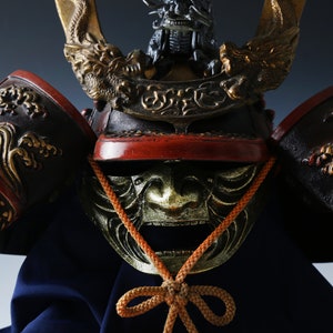 Puede incluir: Primer plano de un casco de samur&aacute;i japon&eacute;s. El casco presenta intrincados dise&ntilde;os dorados de dragones y olas sobre una base negra y roja. Una m&aacute;scara dorada con la boca abierta es visible debajo del casco.