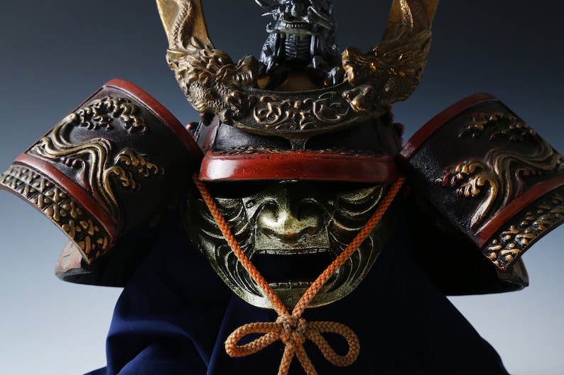 Puede incluir: Primer plano de un casco de samur&aacute;i japon&eacute;s. El casco presenta intrincados dise&ntilde;os dorados de dragones y olas sobre una base negra y roja. Una m&aacute;scara dorada con la boca abierta es visible debajo del casco.