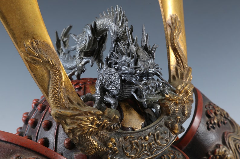 Puede incluir: Primer plano de un casco de samur&aacute;i japon&eacute;s. El casco presenta una pieza central de drag&oacute;n plateado detallada y detalles dorados. El dise&ntilde;o del casco incluye elementos lacados en rojo y negro, mostrando una artesan&iacute;a intrincada.