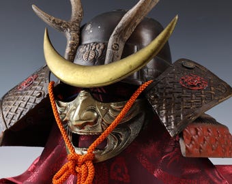 May include: A samurai helmet with a gold crescent moon brow guard, antler accents, and a detailed face mask. The helmet is decorated with intricate patterns and is displayed on a red fabric background.
