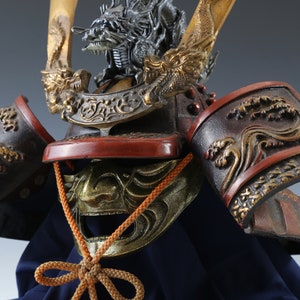Puede incluir: Primer plano de un casco de samur&aacute;i japon&eacute;s. El casco presenta una cresta de drag&oacute;n, intrincados detalles dorados y una combinaci&oacute;n de colores rojo y negro. Los protectores de hombro tienen un dise&ntilde;o de olas.